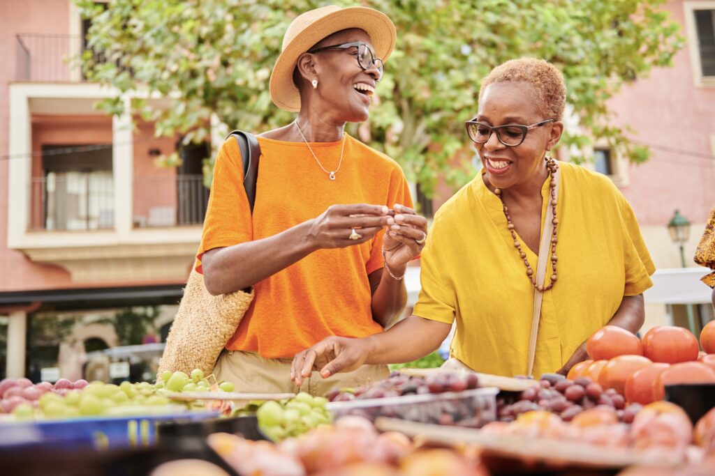 Two mature Black women shop together at an outdoor food market.